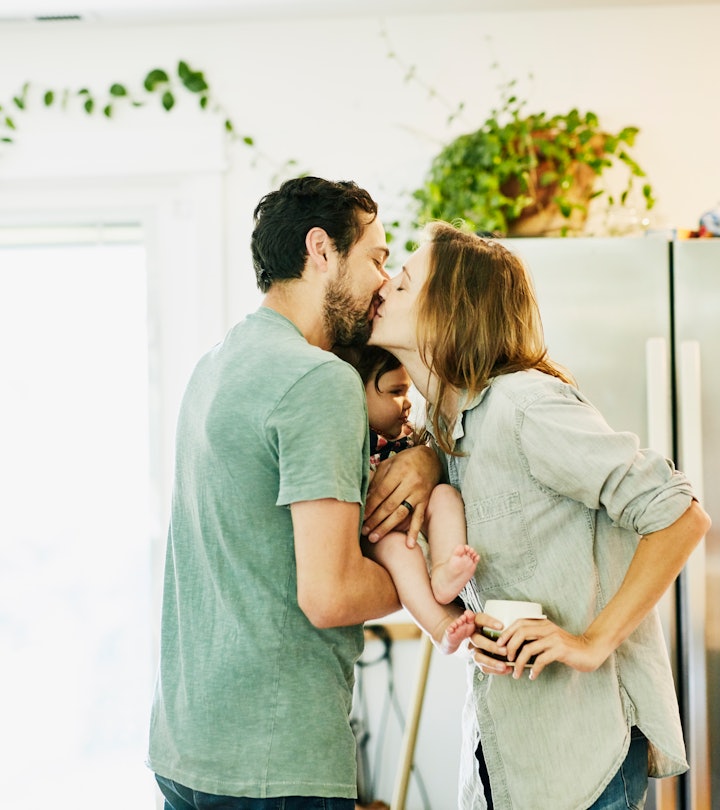 couple sweetly kissing in kitchen, holding infant between them