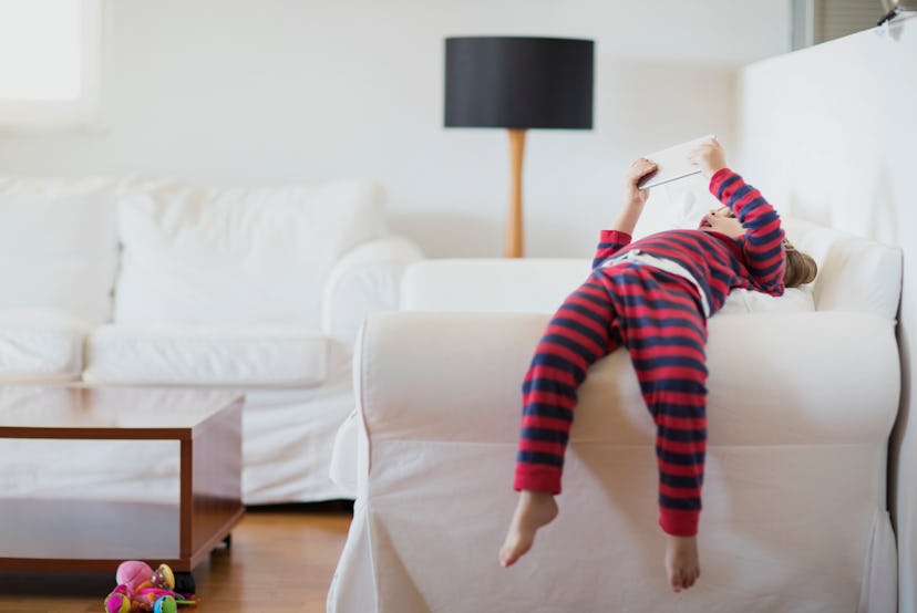 A toddler in striped red and black pajamas lying on the hand rest of a couch and holding a phone abo...