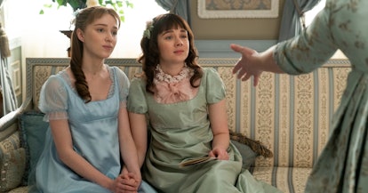 Daphne and Eloise from 'Bridgerton' hold hands in silk dresses while sitting on a couch.