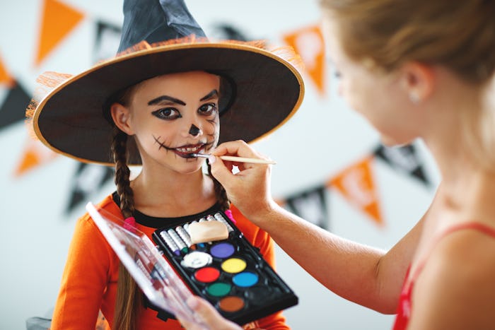 A young woman painting a face of a girl dressed in a witch costume 