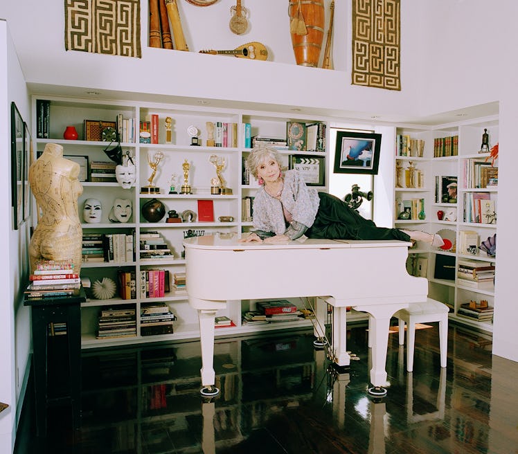 Rita Moreno lies atop a white piano in black pants and gray top surrounded by bookshelves.