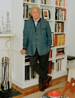 Graydon Carter poses against bookshelf in gray jacket and black pants. 