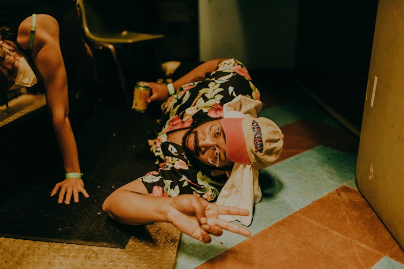 Bartees Strange lying on the floor in a floral shirt and cap while holding a drink