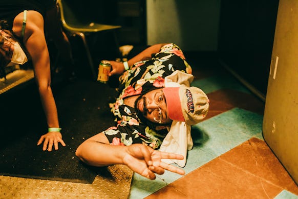 Bartees Strange lying on the floor in a floral shirt and cap while holding a drink
