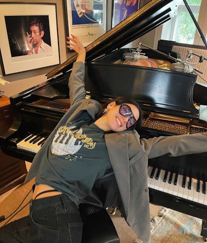 Dua Lipa wearing an Elton John shirt while leaning against a piano