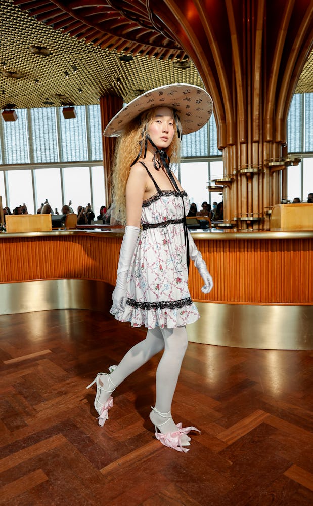 A model wearing a white-black floral dress, white gloves, white tights and sandals and a hat by Mais...