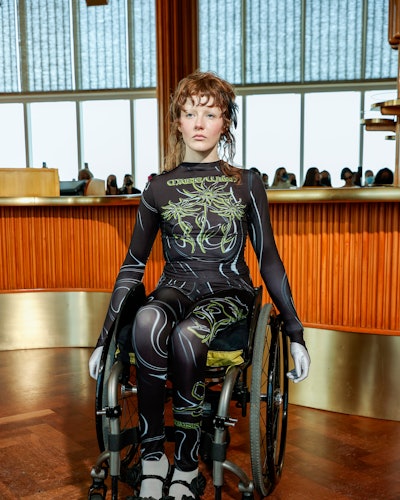 A model in a wheelchair wearing a tulle top and leggings in black with white and yellow print by Mai...