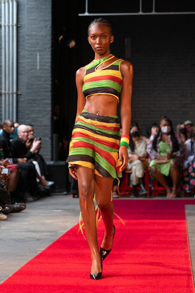 A model wearing a matching striped top and skirt in black, yellow, orange, and red by Theophilio