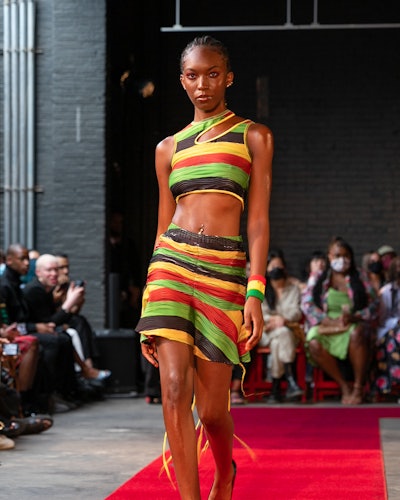 A model wearing a matching striped top and skirt in black, yellow, orange, and red by Theophilio
