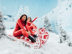 Two women sit in sleighs from FUNBOY's Winter 2021 collection on a snowy mountain.