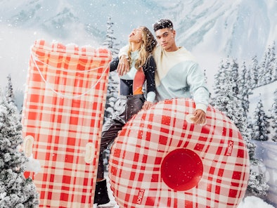 A couple stands on a snowy mountain with tubes from FUNBOY's winter 2021 collection.