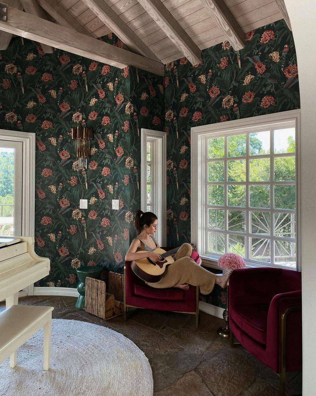 Selena Gomez sitting in a chair playing a guitar in her room with a floral wallpaper