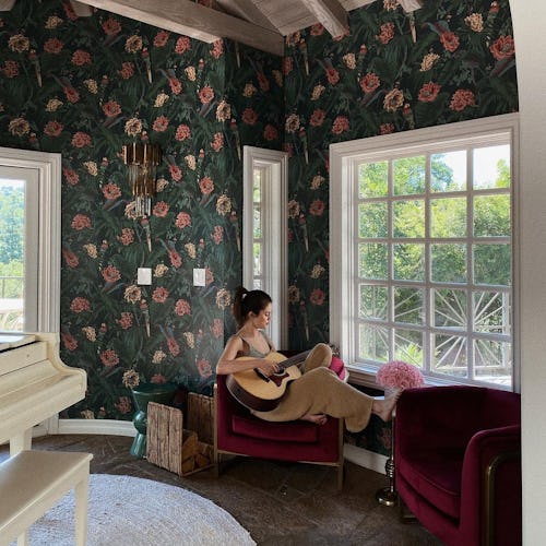 Selena Gomez sitting in a chair playing a guitar in her room with a floral wallpaper
