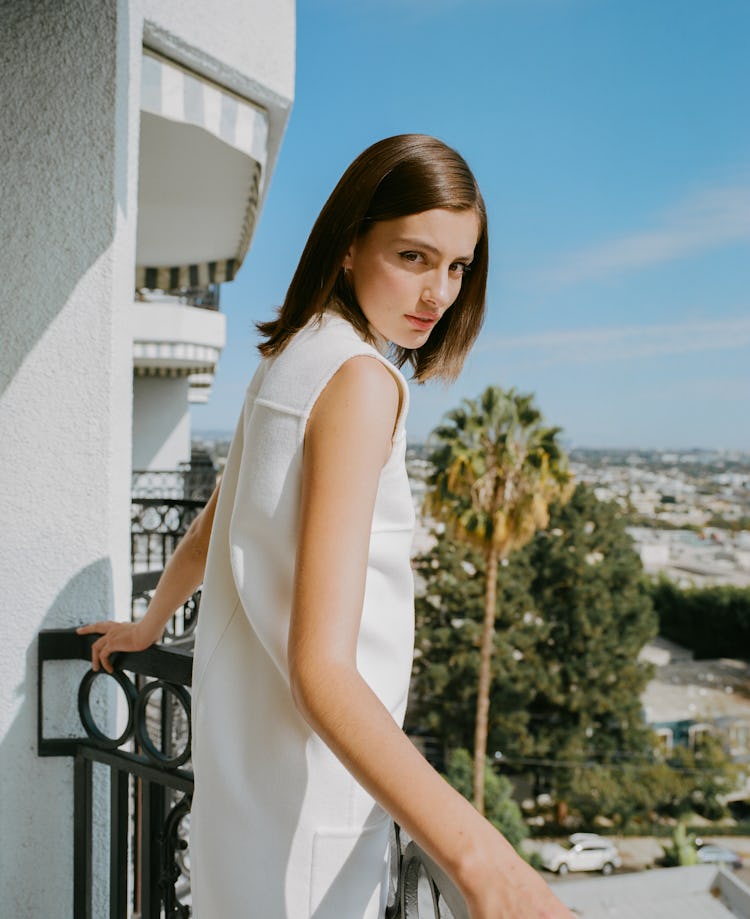 Diana Silvers posing for a picture in white dress at the edge of a balcony with a sea view.