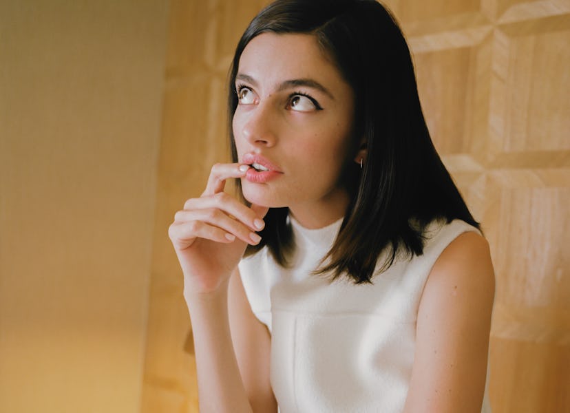 Diana Silvers posing for a picture in white dress while looking up and keeping her finger on the lip...