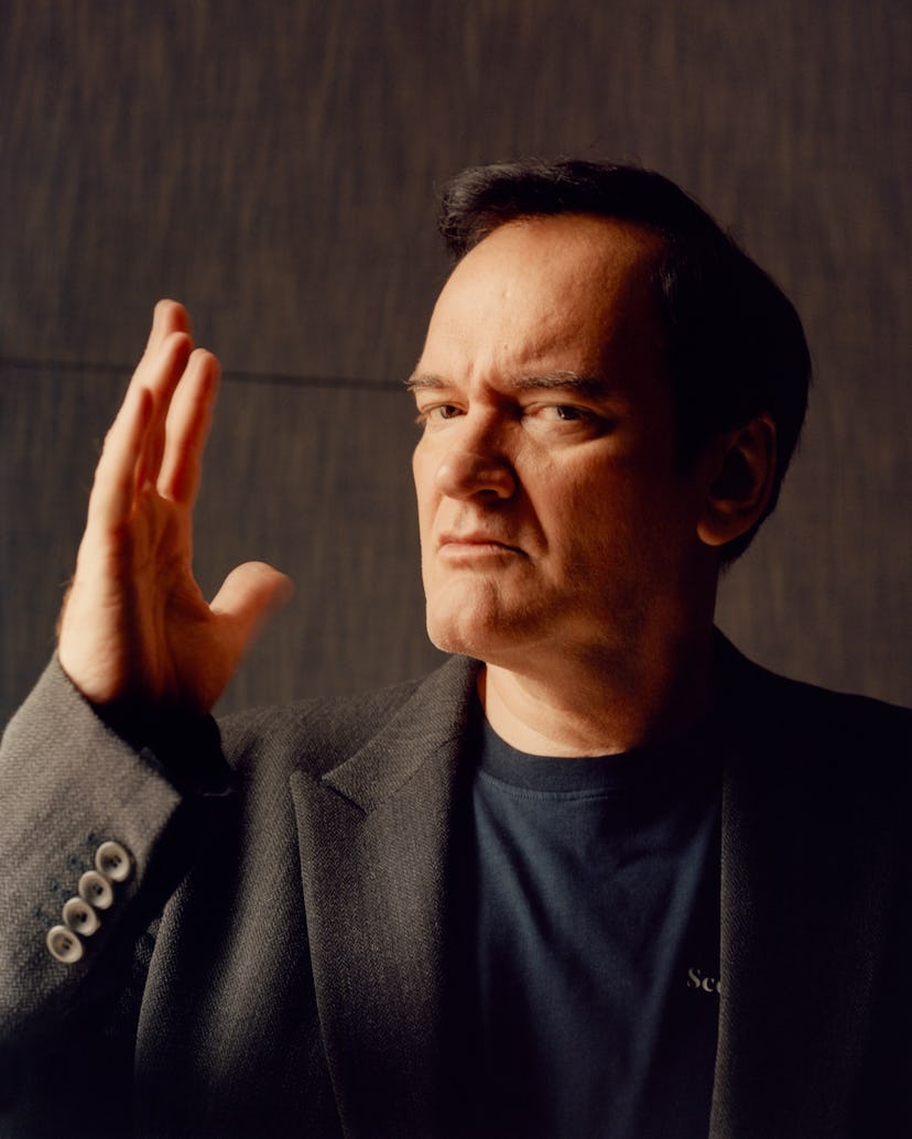 A portrait of Quentin Tarantino in a black shirt and a brown blazer with one hand up