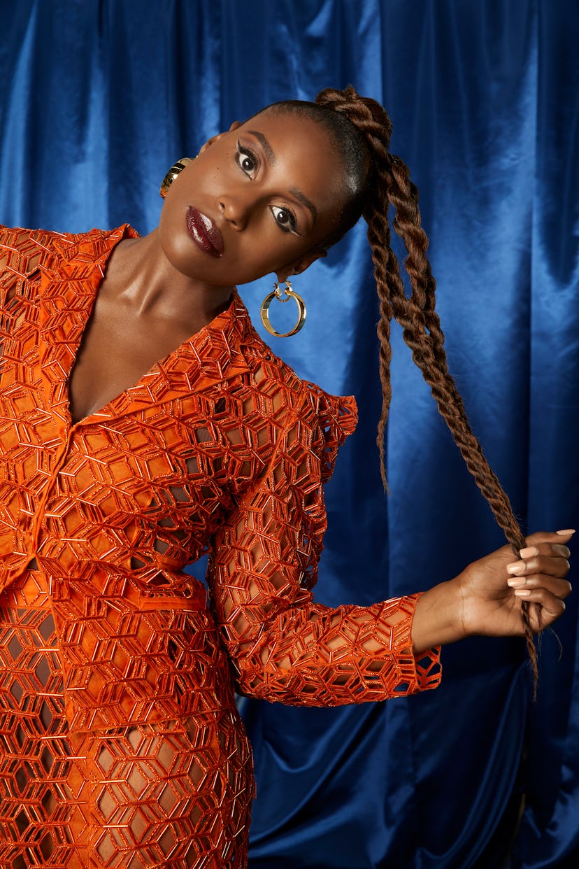 Issa Rae in an orange Aliette suit and Sister Love jewelry looking to the camera, pulling her long b...