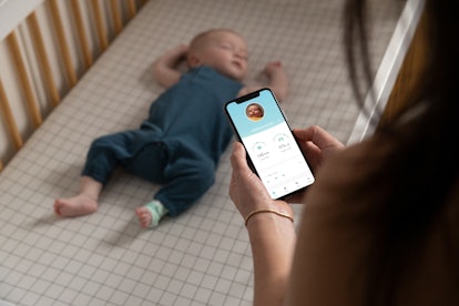 A woman looks at the Owlet app on a smartphone while a baby lies on its back in a crib while wearing...