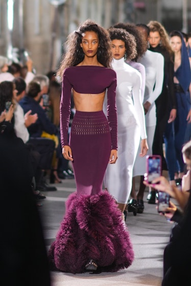 A model wears a purple long sleeves crop-top, a purple tube long skirt with fluffy feathers on the a...