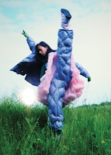 a model wearing a puffer jacket, pants and a tutu does a high kick in a grassy field