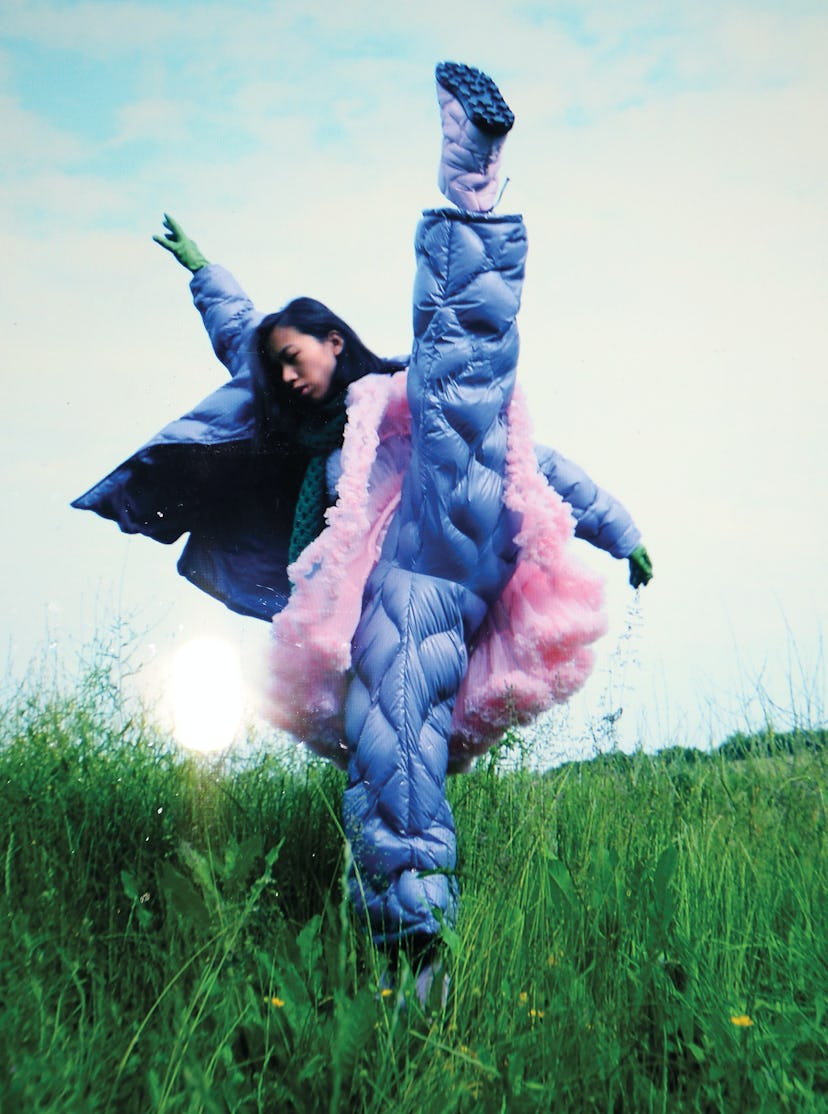 a model wearing a puffer jacket, pants and a tutu does a high kick in a grassy field