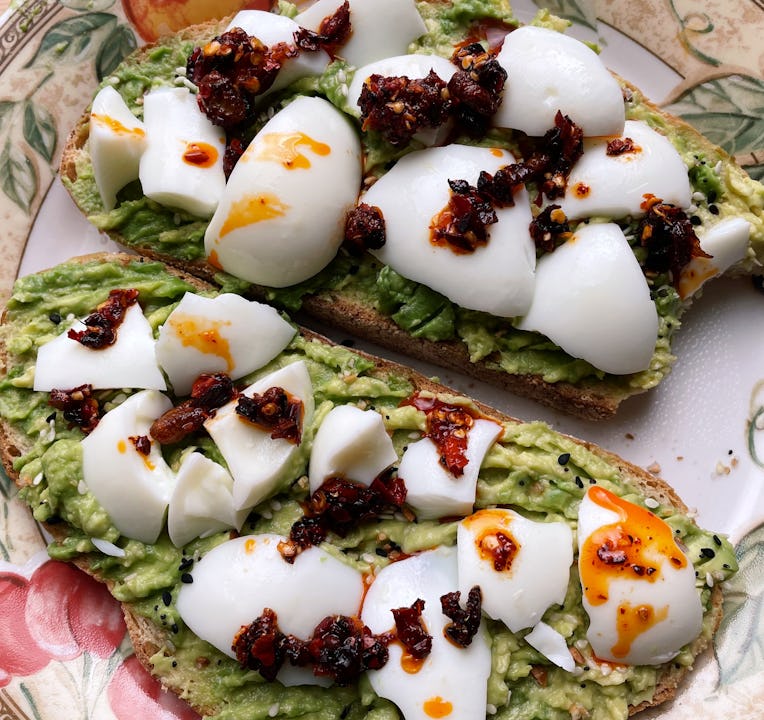 Close-up of two slices of toast spread with avocado, chunks of hard-boiled egg, and chili oil.