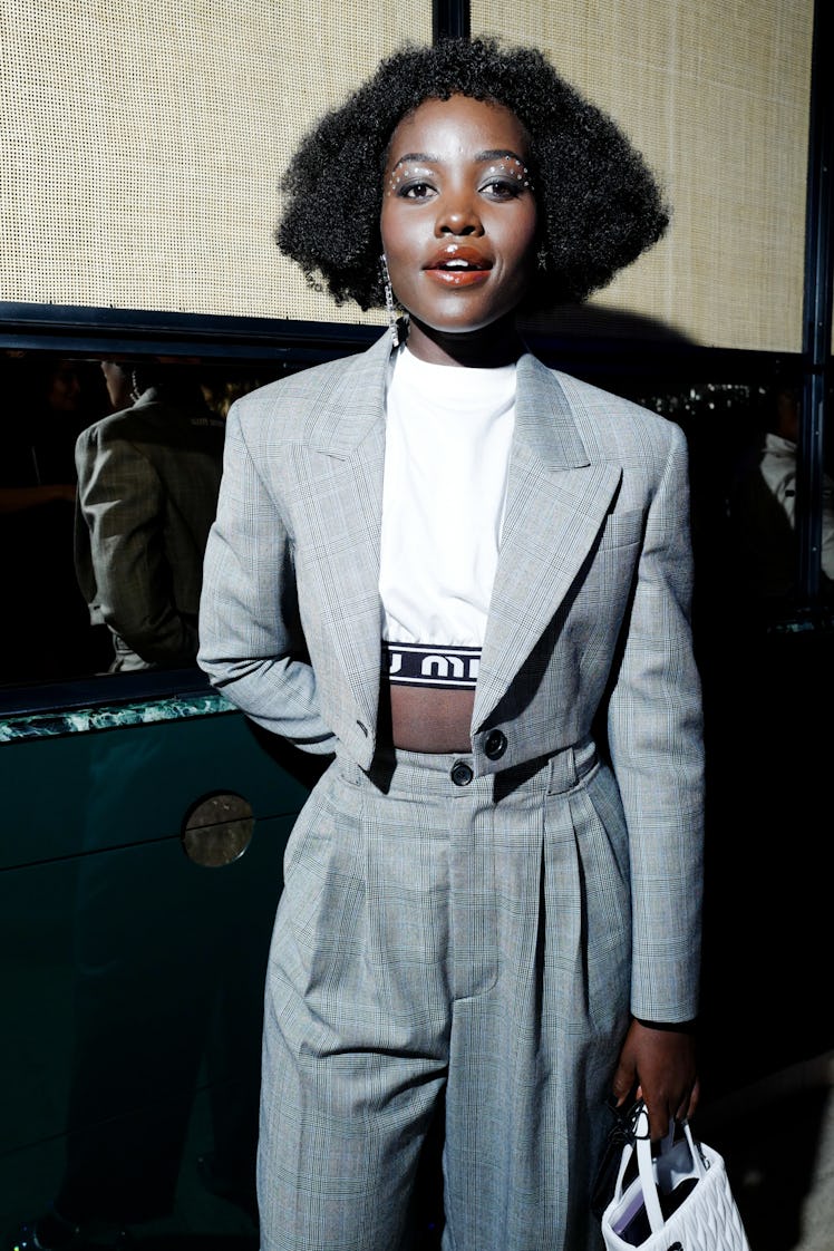 Lupita Nyong'o posing in a small grey blazer, white top, and grey pants