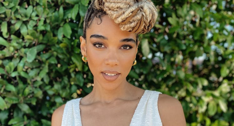 A portrait of Alexandra Shipp in a black-white dress with a bow posing in front of a green bush