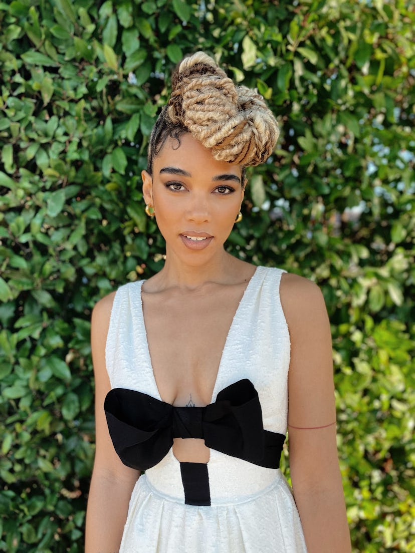 A portrait of Alexandra Shipp in a black-white dress with a bow posing in front of a green bush