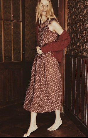 A woman wearing a red printed cotton dress and cardigan sweater with care feet in a house entryway.