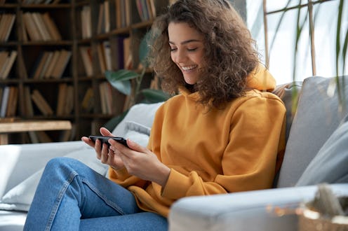 A young woman in a yellow sweatshirt looks down at her phone and smiles. "Add yours" sticker not wor...