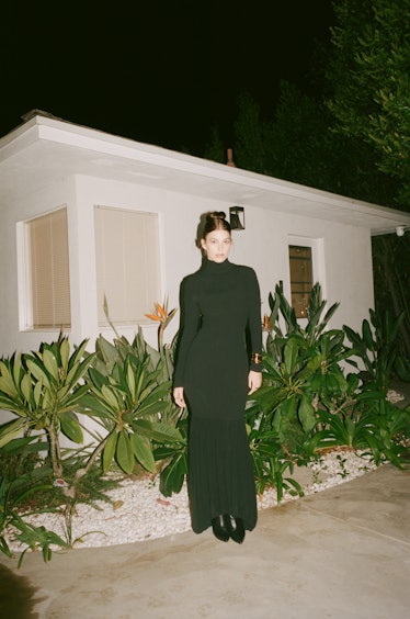 Camilla Morrone wearing a long black dress standing outside of a modern house at night