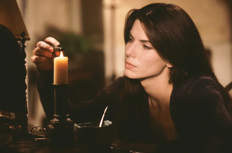 A women with white skin and brown hair holds something over a lit candle. She has a focused expression, and is wearing a dark sweater and sitting at a desk.
