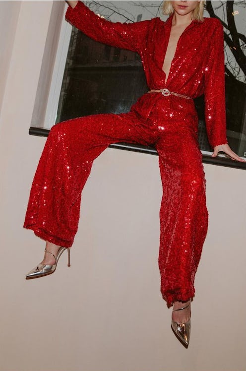A model sitting on a window ledge wearing a red sequin jumpsuit and crystal-embellished pumps.