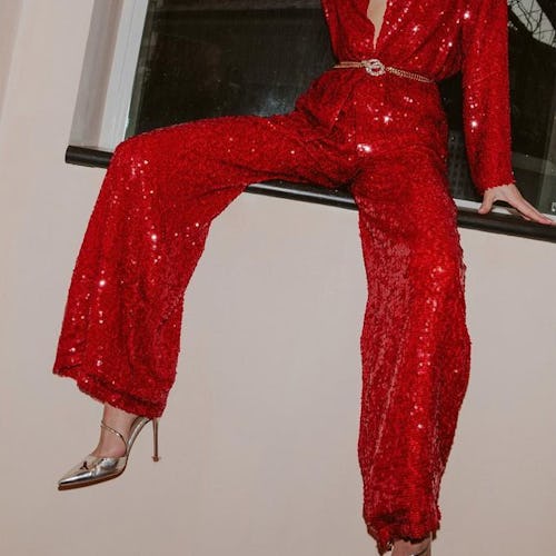 A model sitting on a window ledge wearing a red sequin jumpsuit and crystal-embellished pumps.