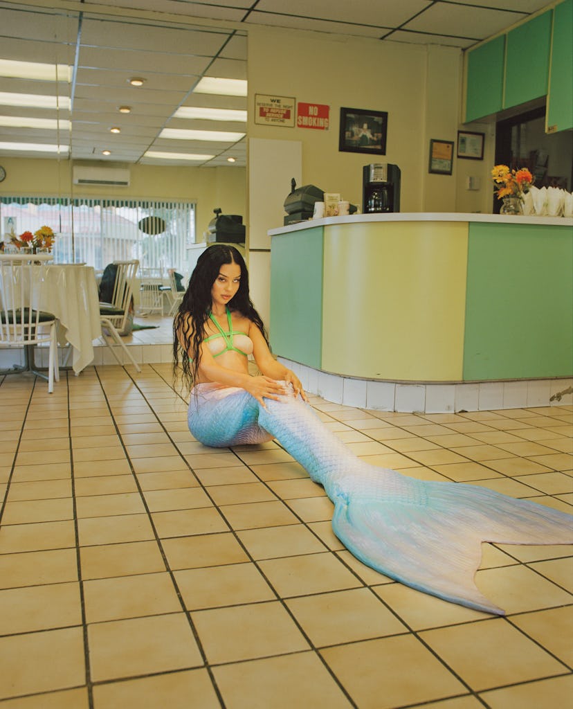 Alexa Demie sitting on a floor of a restaurant, wearing a mermaid outfit with one white eye contact