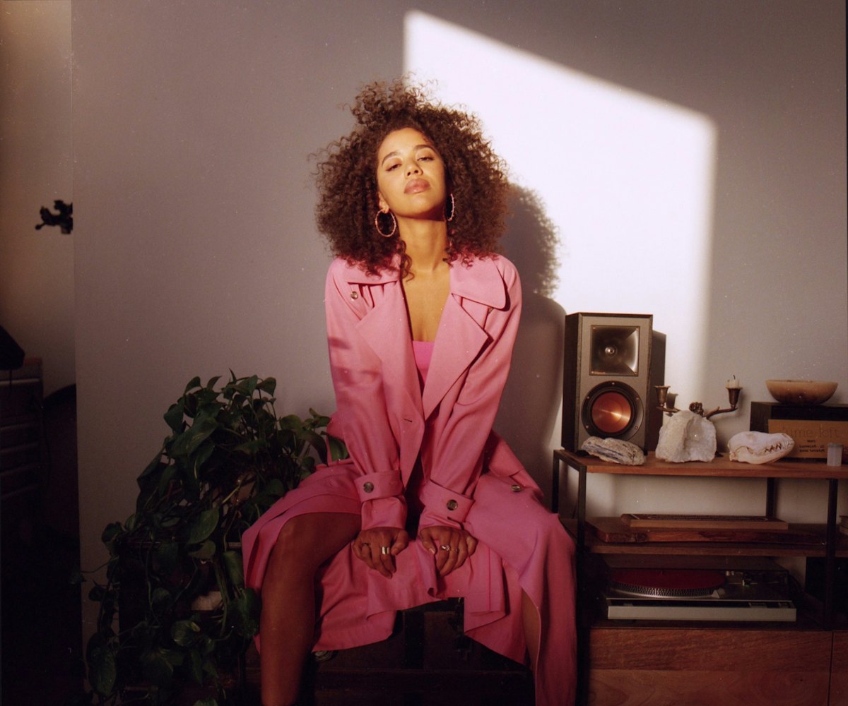 Jasmin Savoy Brown is wearing in a pink dress and a long pink trench-coat, curly hair, and large hoo...