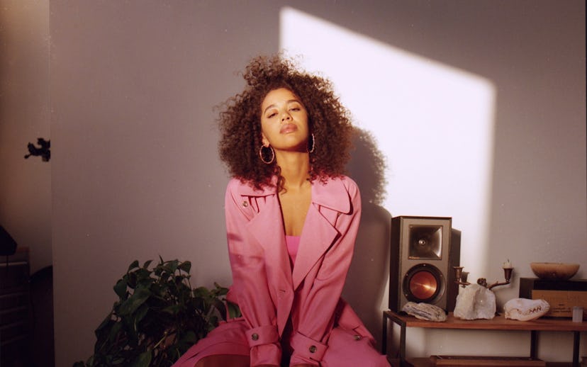 Jasmin Savoy Brown is wearing in a pink dress and a long pink trench-coat, curly hair, and large hoo...
