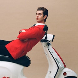 Andrew Garfield in a red blazer, black pants and a white button-up lying on top of a Vespa