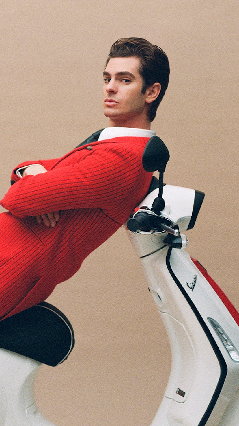 Andrew Garfield in a red blazer, black pants and a white button-up lying on top of a Vespa
