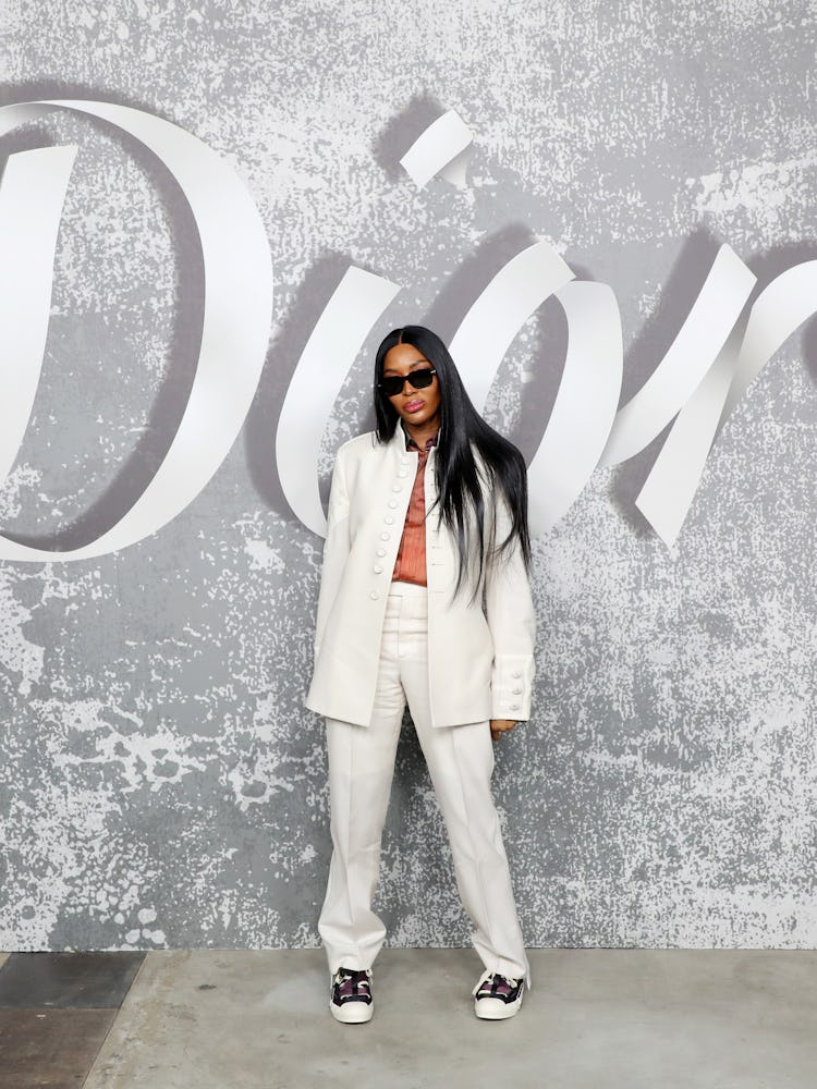 Naomi Campbell in a white suit and black sneakers with a Dior background