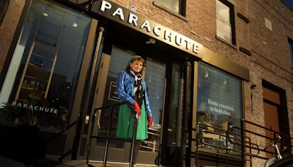 Norah Jones in a green beret and skirt, red gloves and a blue coat in front of a parachute store