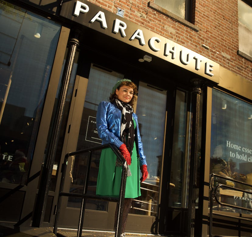 Norah Jones in a green beret and skirt, red gloves and a blue coat in front of a parachute store