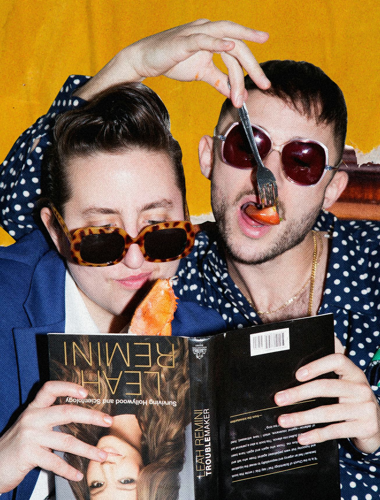 Two people reading Leah Reminis book upside down while wearing sunglasses and eating fruit with a fo...