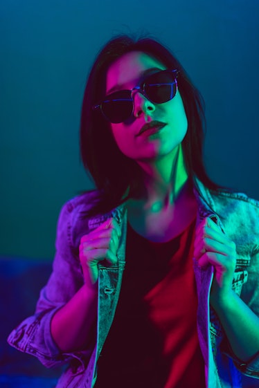 Young woman with sunglasses in neon lights before reading her December 20, 2021 weekly horoscope for...