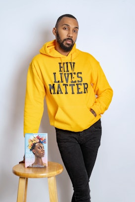 George M. Johnson standing in front of a table with his book on it, in a yellow sweatshirt that says...