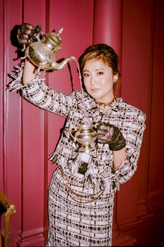 Ashley Park in a matching black-and-white tweed jacket and skirt, sipping tea from a golden teapot i...