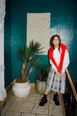 Ashley Park in a white shirt, red sweater over her shoulder, a grey check skirt and black sneakers