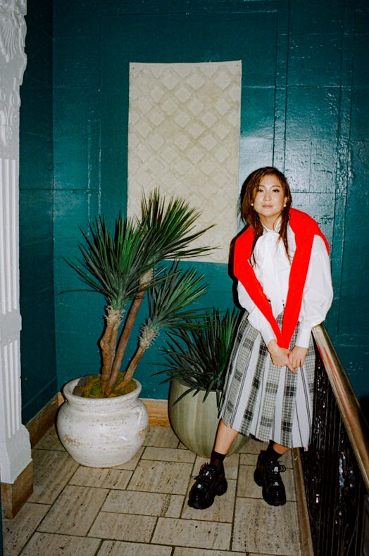 Ashley Park in a white shirt, red sweater over her shoulder, a grey check skirt and black sneakers