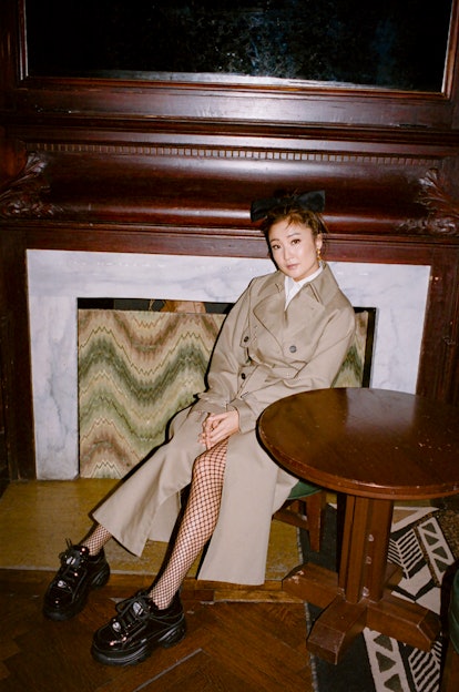 Ashley Park sitting and posing in a beige trench coat, fishnet tights and black sneakers